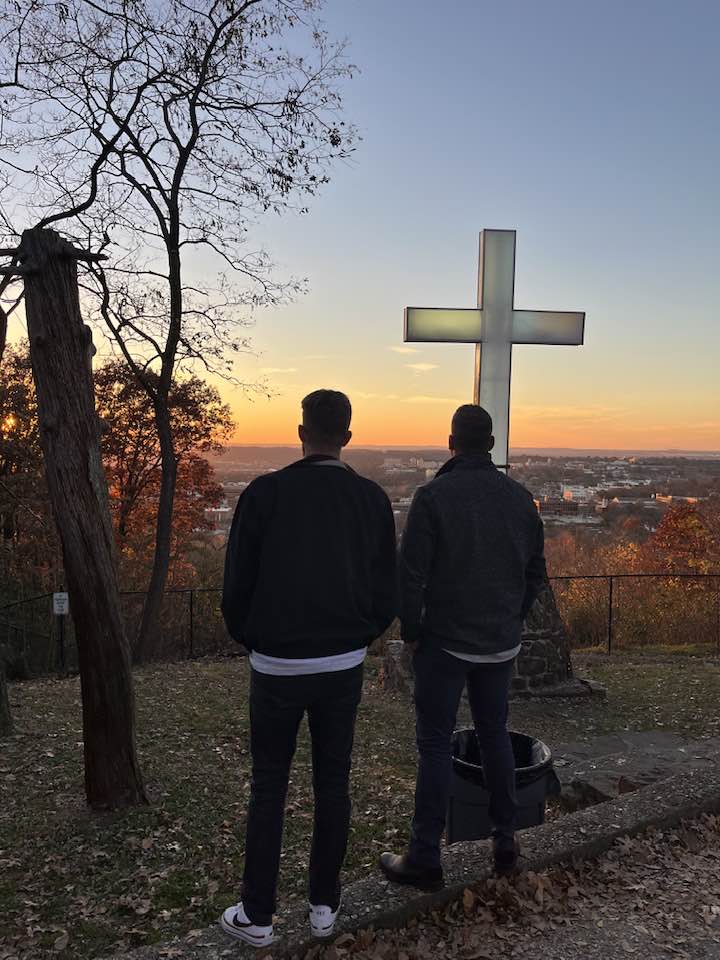 Two men in contemplation before an illuminated cross at sunset, symbolizing faith and spiritual reflection in recovery